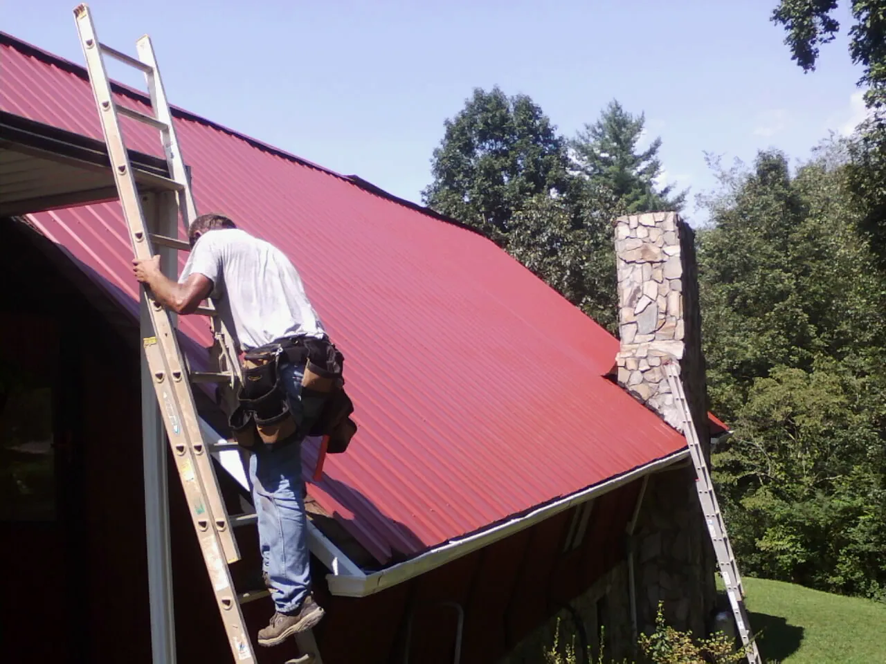 Roofer on a ladder beside a red metal roof and stone chimney — Soffit & Fascia Repair by Roofing Brothers in Guys